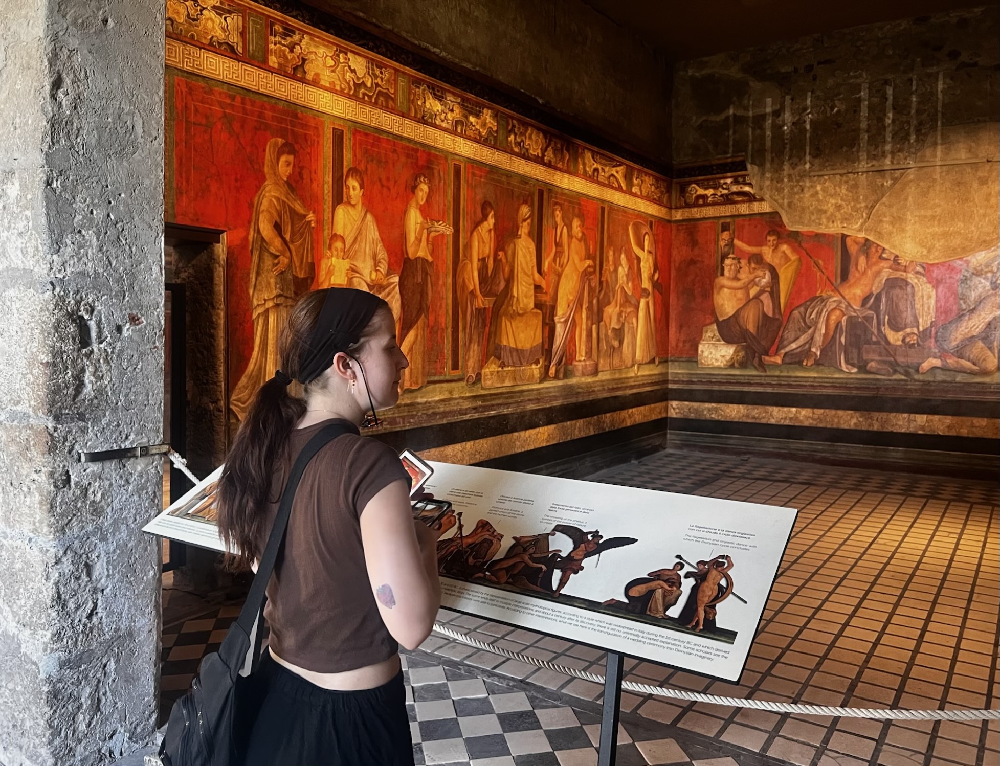 A young woman stands inside an ancient room in Pompeii, observing a vivid red fresco depicting mythological scenes, with an informational display in front of her.