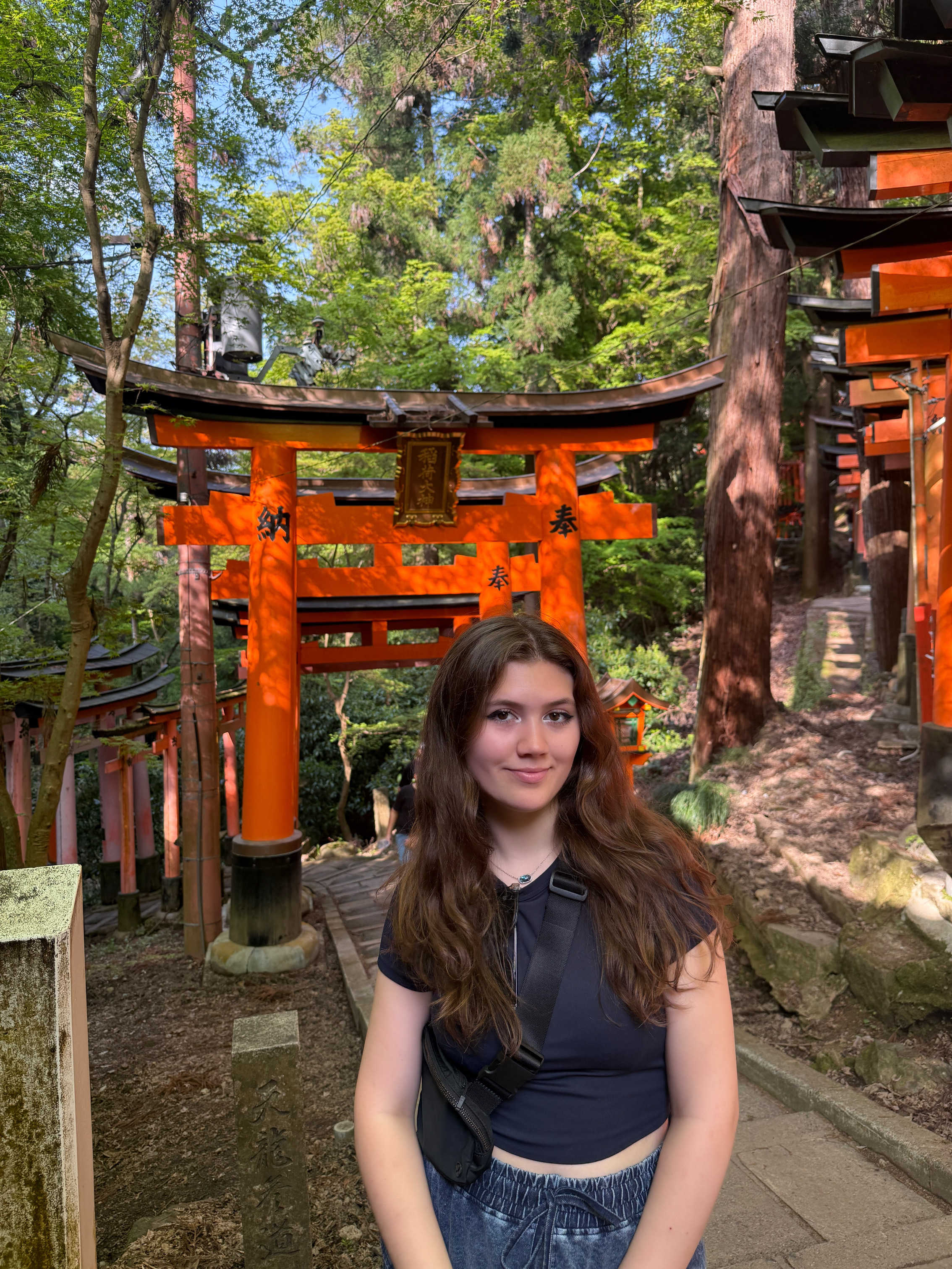 A young woman stands smiling at a wooded shrine path lined with vibrant orange torii gates in Japan.