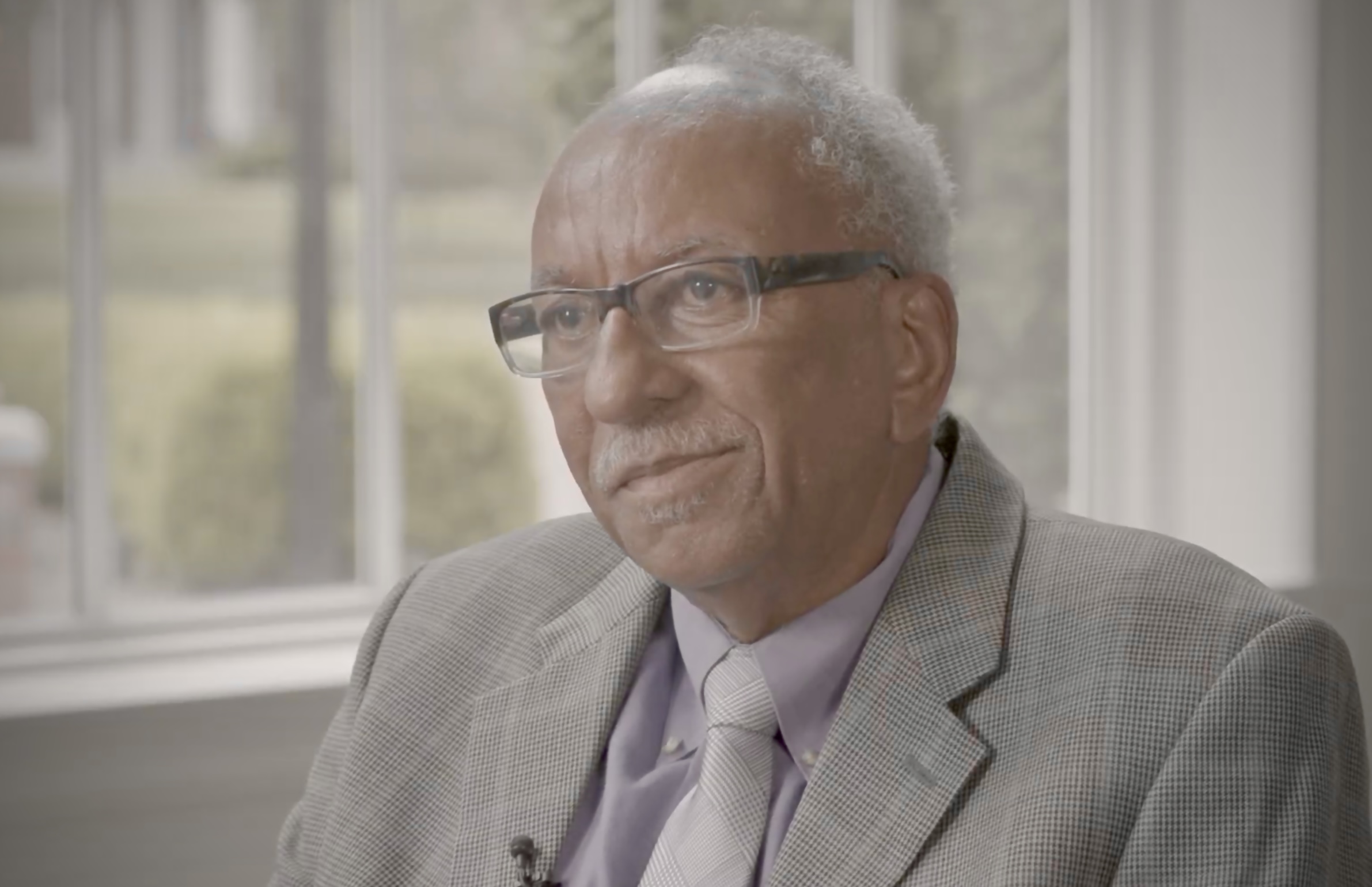 A man in a gray suit jacket, lavender shirt, and glasses sits in front of a bright window, looking thoughtful.