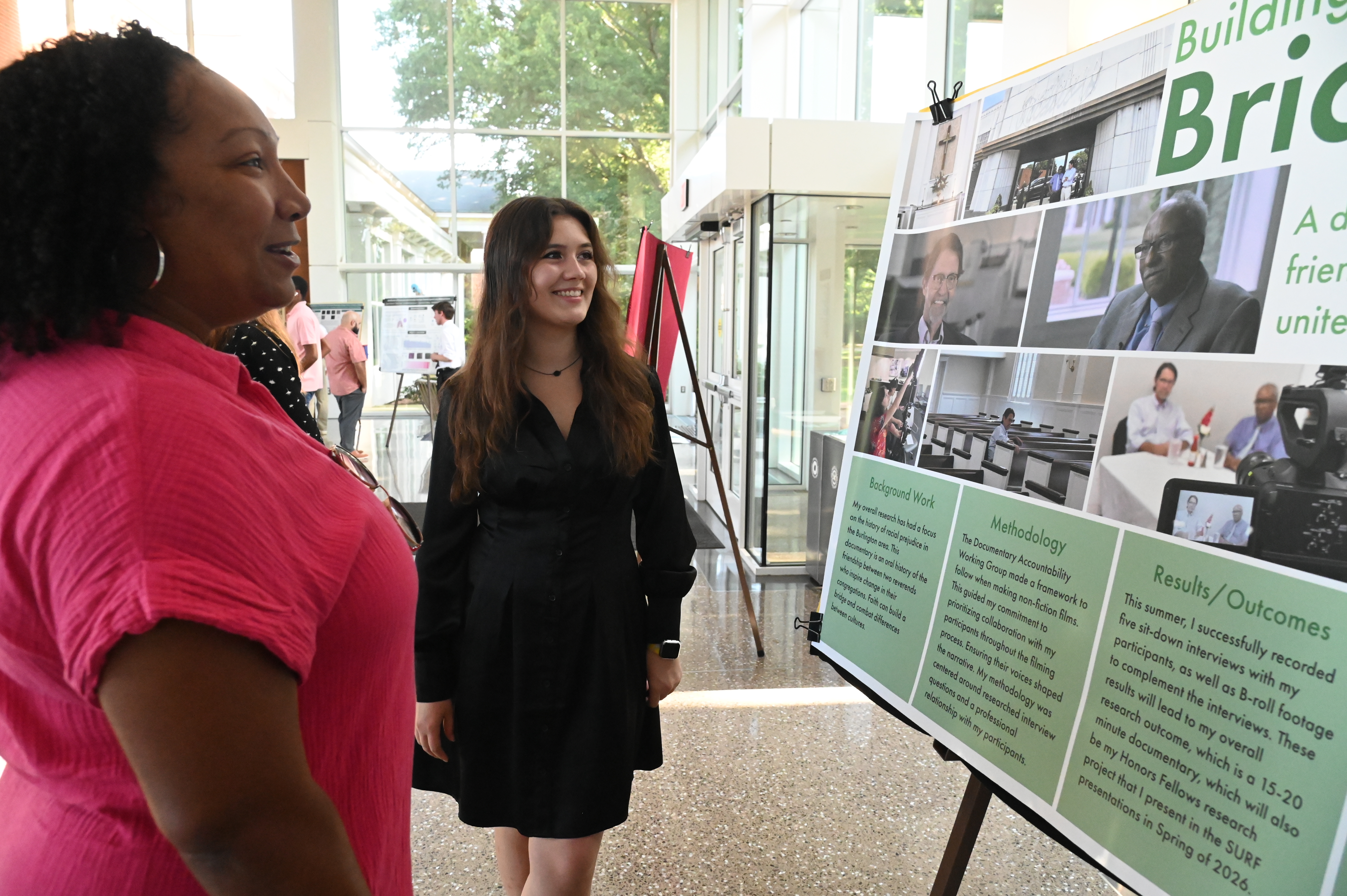 A student in a black dress discusses her research poster with an attendee during an academic showcase in a bright, glass-walled atrium.