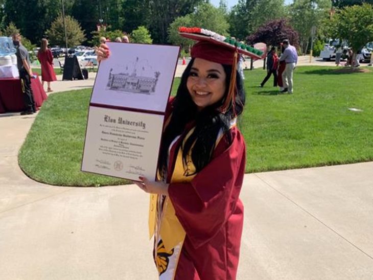 A woman in graduation regalia smiles as she holds up her diploma from Elon University