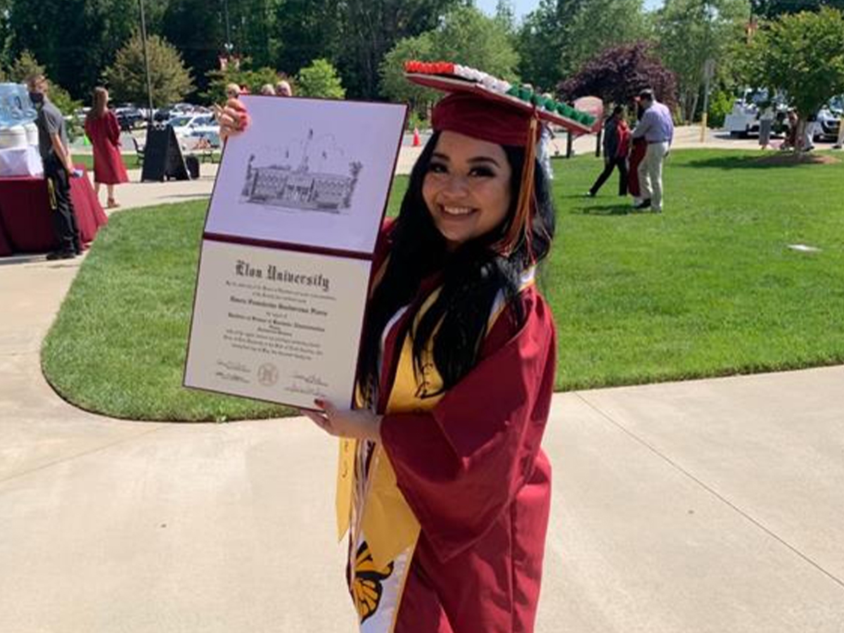 A woman in graduation regalia smiles as she holds up her diploma from Elon University