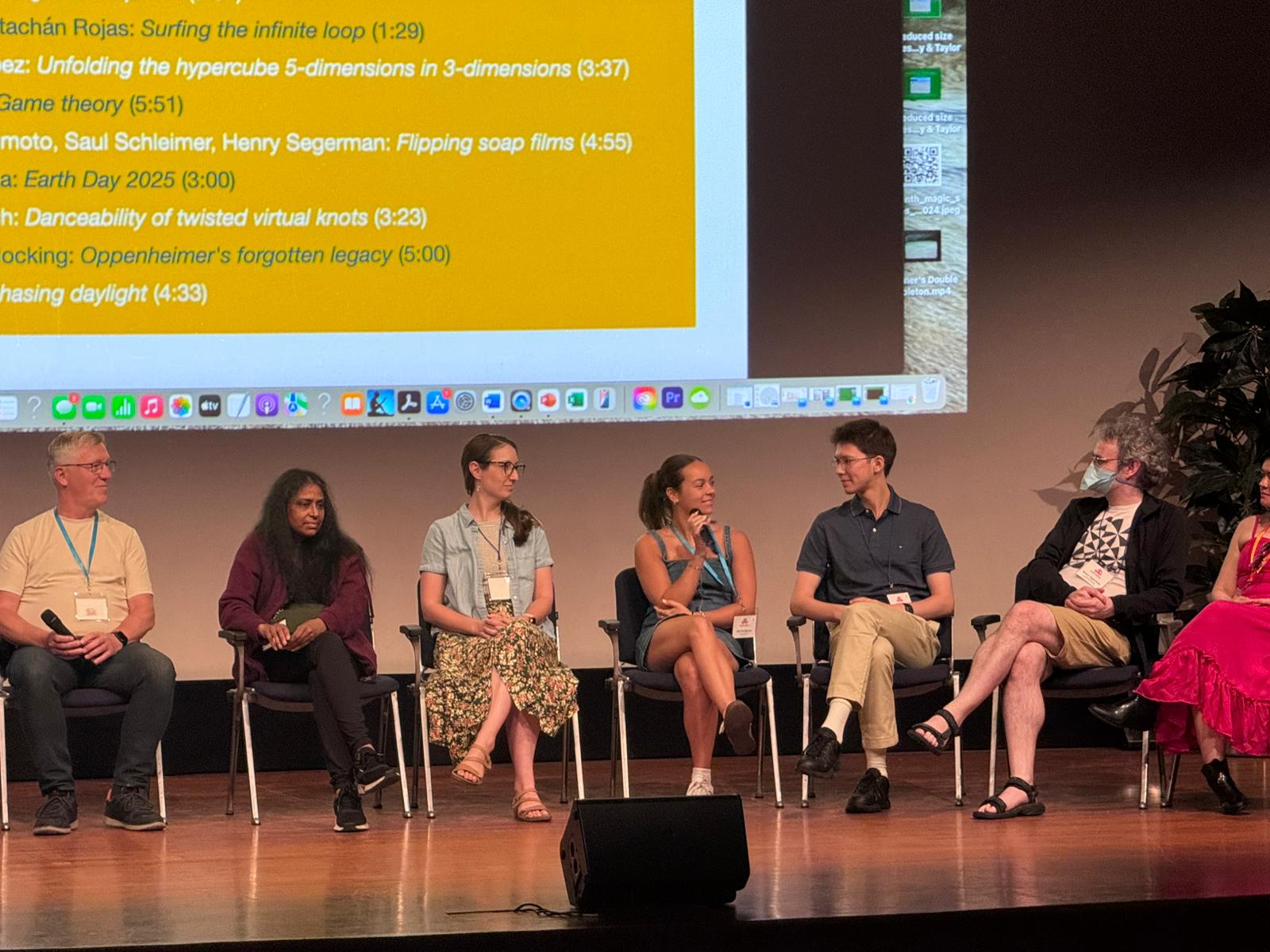 A group of seven panelists sit on stage in front of a projected screen, engaged in discussion during what appears to be a conference or academic event.