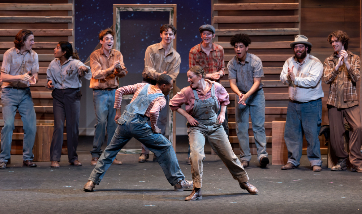 Two performers tap dance at center stage while cast members in rustic costumes cheer them on during a theatrical production.
