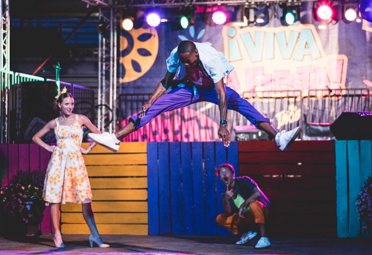A performer leaps into a mid-air split on stage during a colorful outdoor show, while two others react with excitement below and to the side.