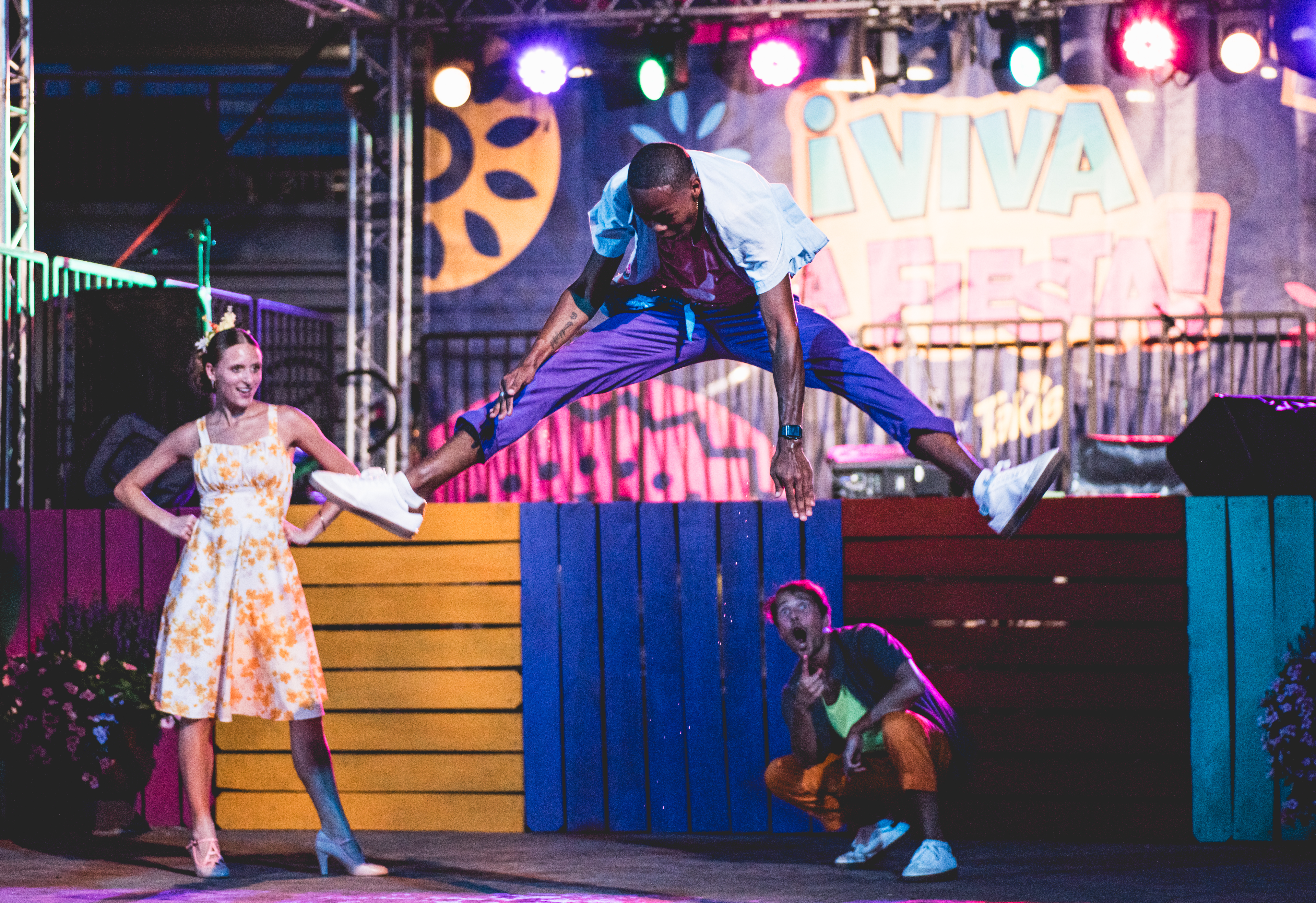 A performer leaps into a mid-air split on stage during a colorful outdoor show, while two others react with excitement below and to the side.