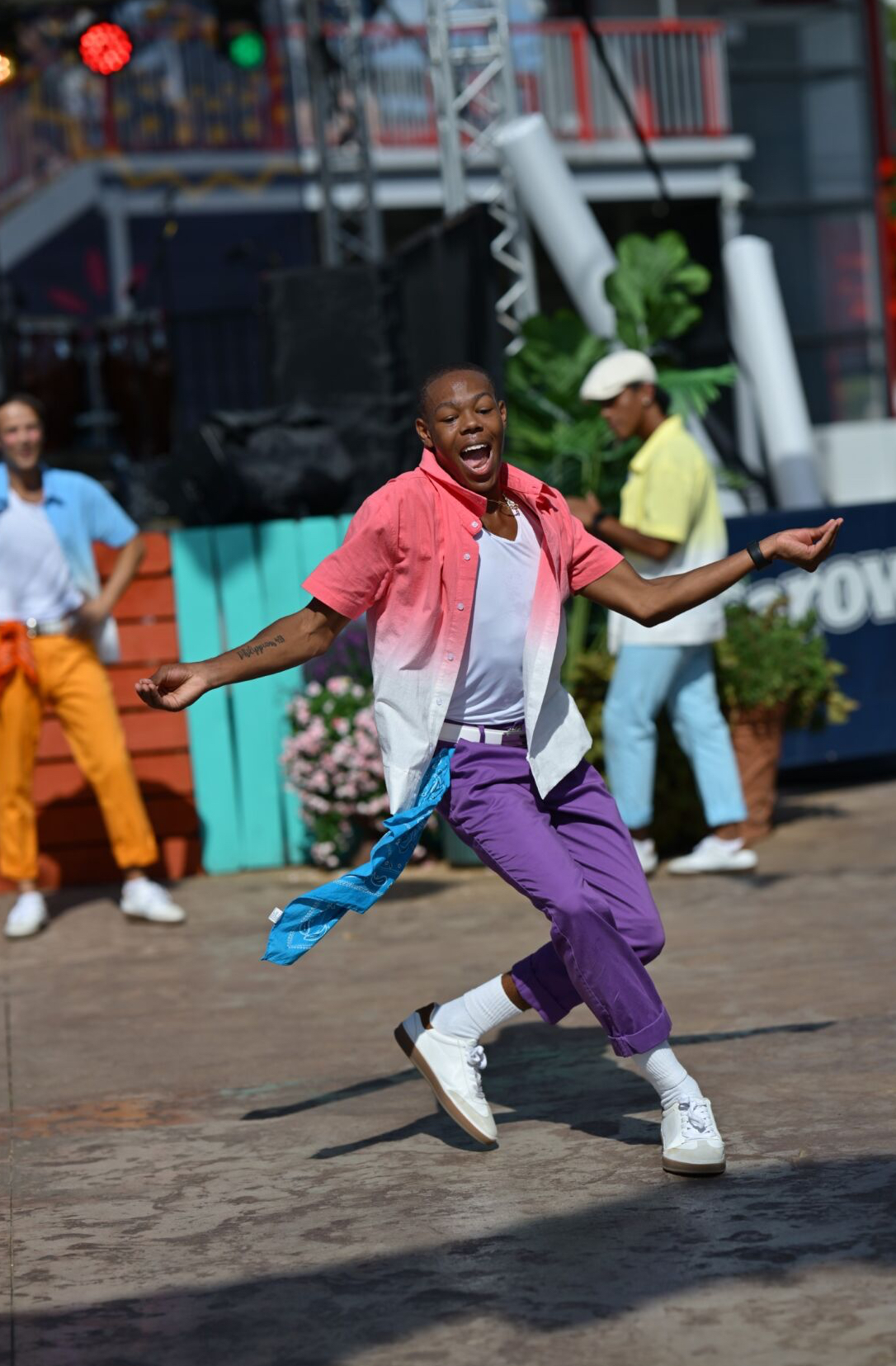 A performer in colorful clothing dances energetically outdoors, with two background dancers and a bright stage setup behind him.