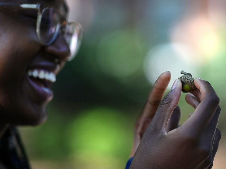 A person smiles while closely examining a small acorn held between their fingers.