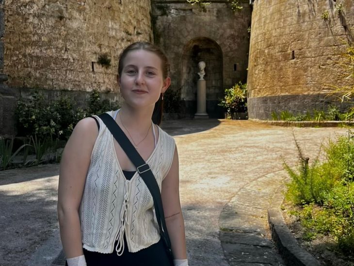 A young woman stands in front of ancient stone walls and greenery at the entrance to Vergil’s Tomb in Naples, Italy, holding a phone and wearing a light knit top and black skirt.