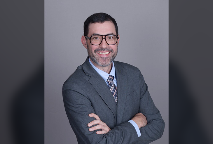 Jonathan Perry L'10 in a gray suit and tie. He is wearing glasses and has his arms crossed, smiling at the camera.