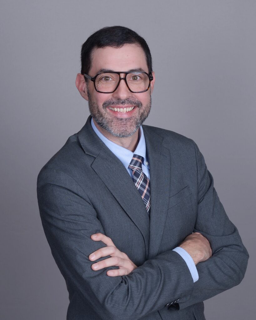 Jonathan Perry L'10 wearing a gray suit, tie and glasses. He has his arms crossed and is smiling at the camera.