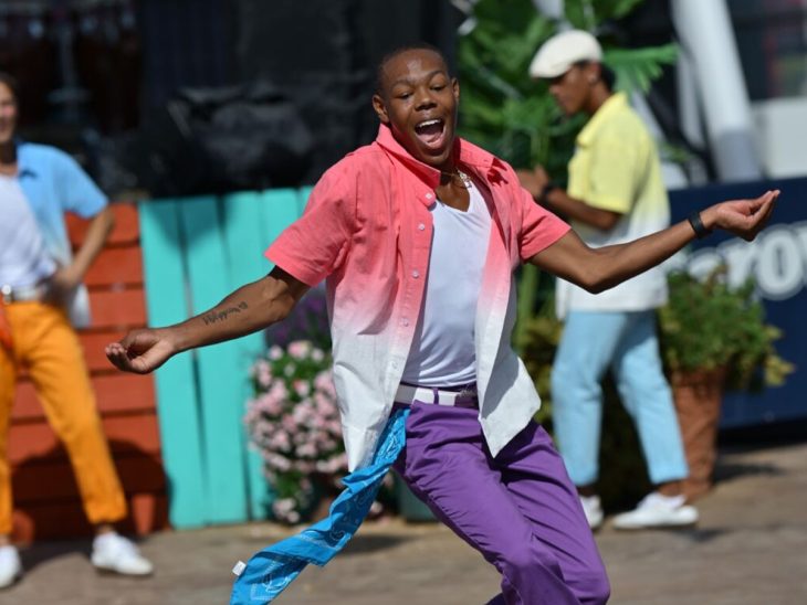 A performer in colorful clothing dances energetically outdoors, with two background dancers and a bright stage setup behind him.