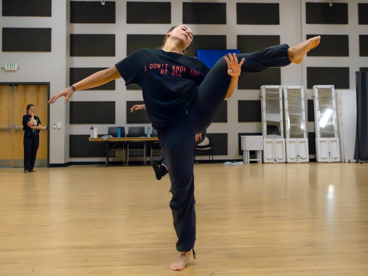 A dancer balances gracefully on one foot during a rehearsal, with an instructor observing in the background.