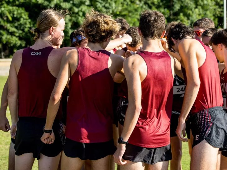 A group of male cross-country runners in matching maroon uniforms huddle together in a pre-race team circle.