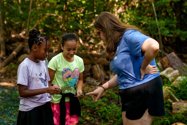 Ellie Loewenthal shows two young girls leaves during an outdoor nature activity in the woods.