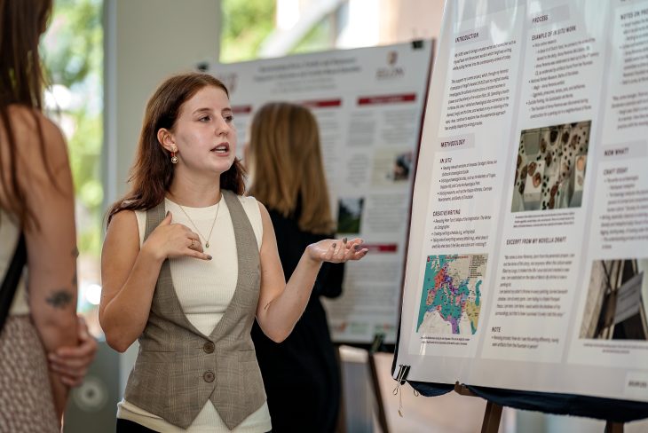 A student gestures while presenting her research poster to attendees during an academic showcase.