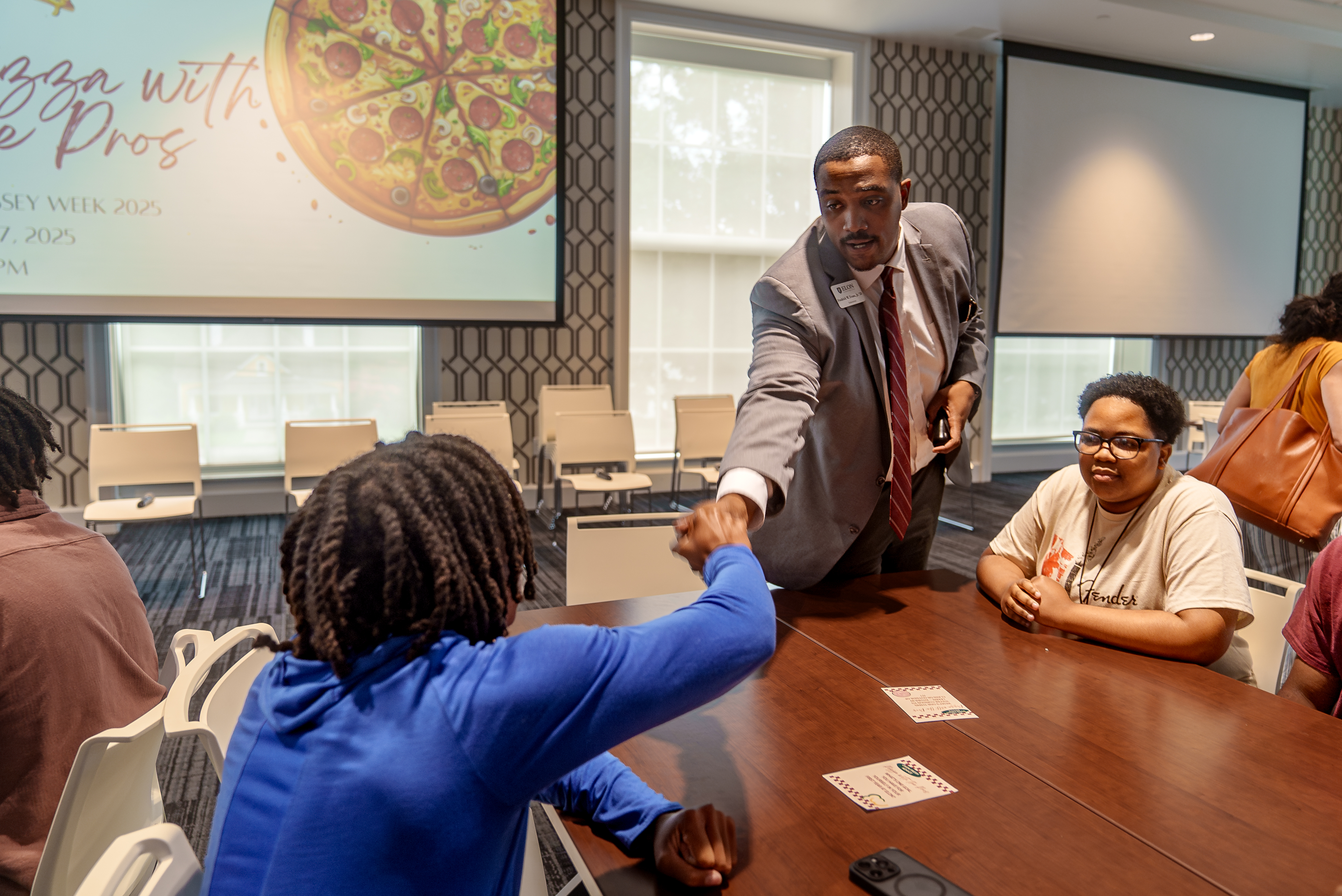 A professional in a suit shakes hands with a student during the “Pizza with the Pros” Odyssey Week 2025 event.