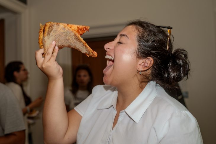 A smiling person prepares to take a big bite of a slice of pizza at an indoor event.