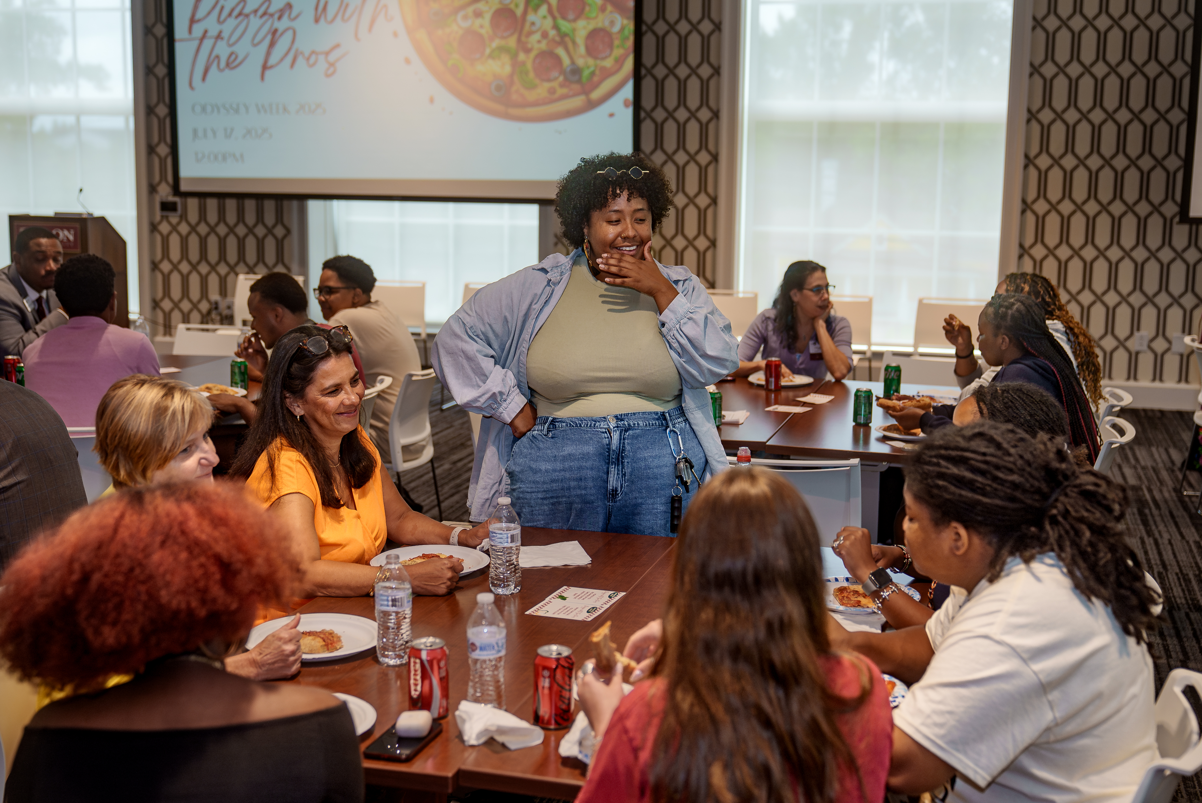 A smiling participant stands and chats with attendees seated around a table during the “Pizza with the Pros” Odyssey Week event.