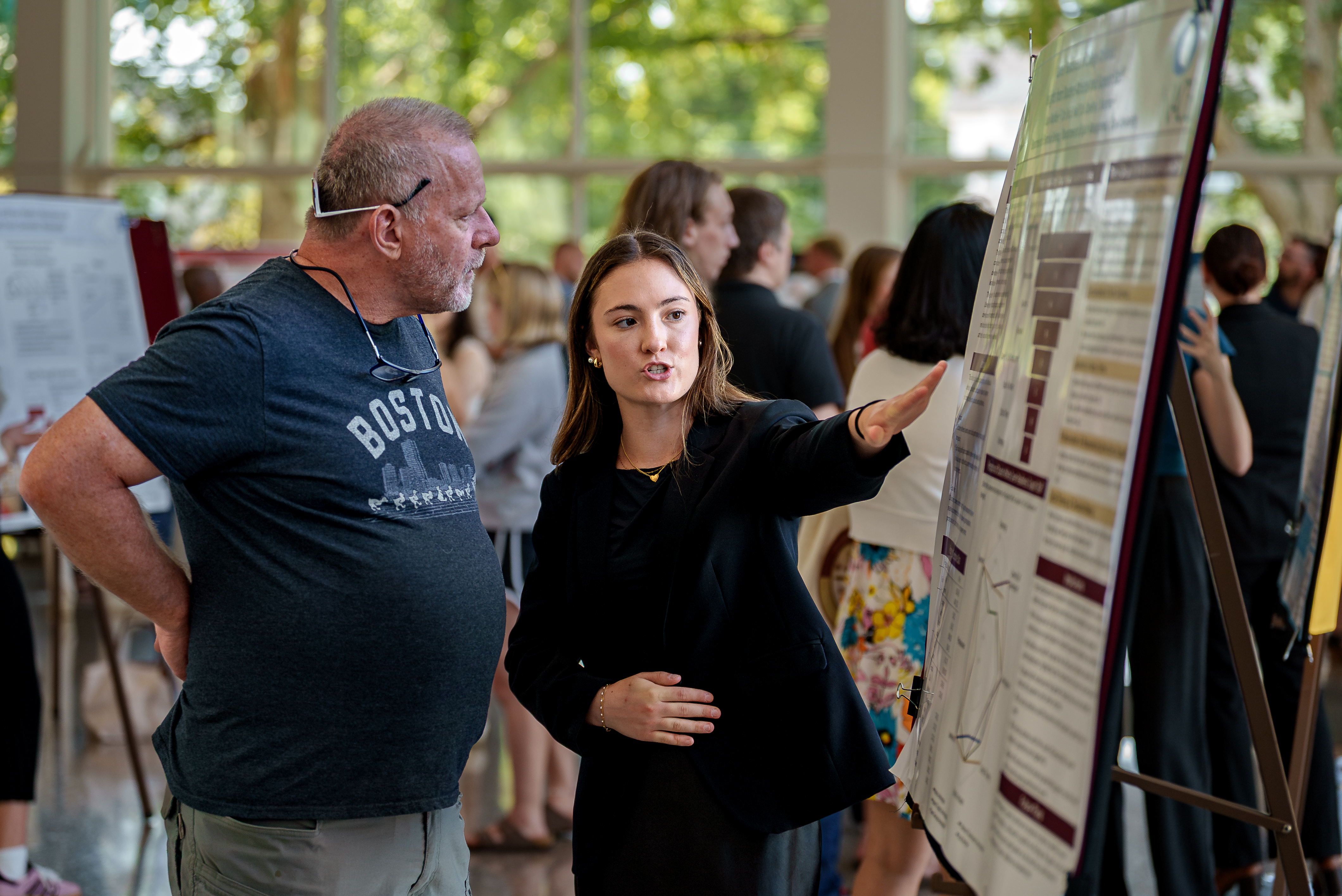 A student in a black blazer points to a section of her research poster while explaining it to an attendee during a busy academic showcase.