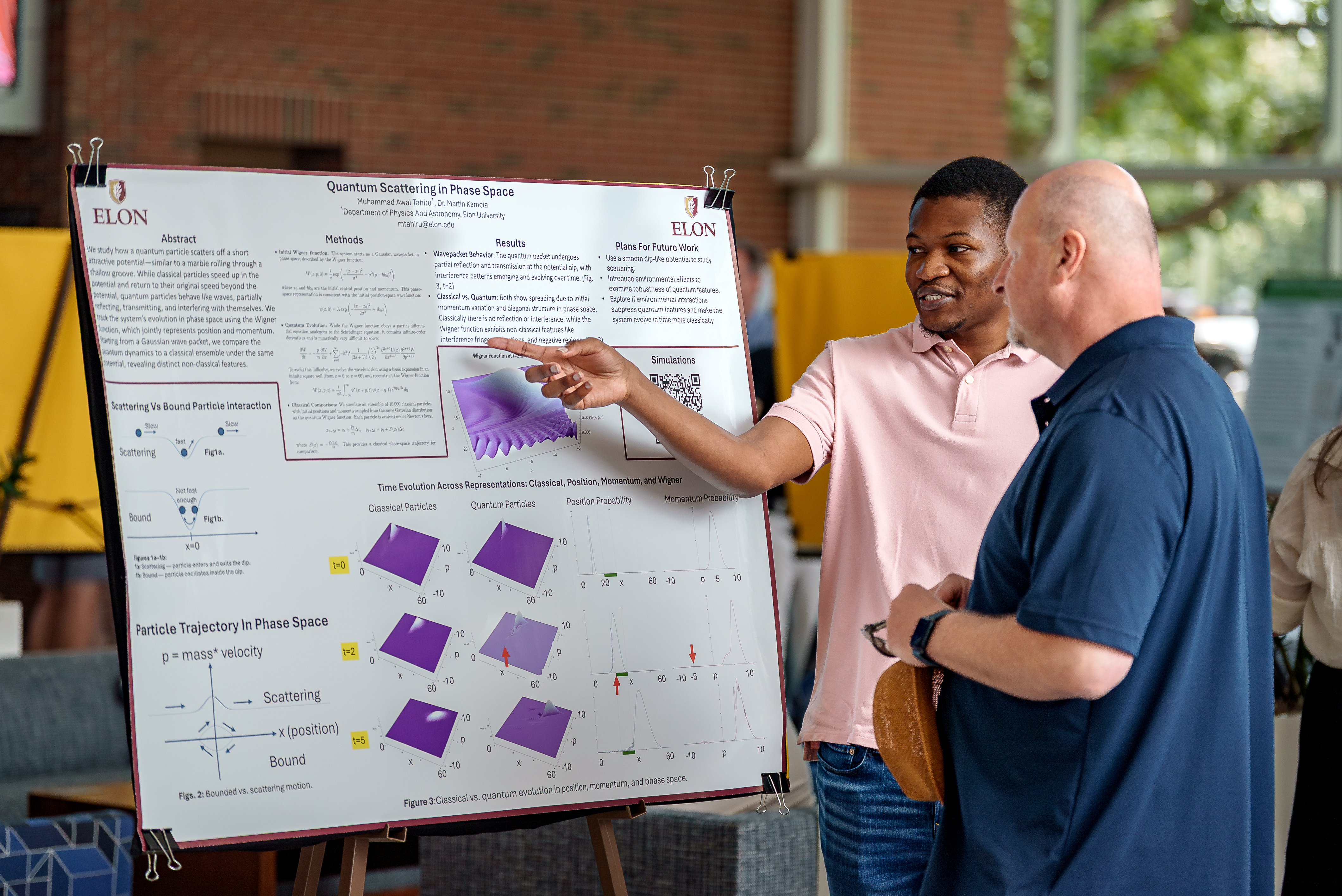 A student in a pink shirt explains a physics research poster featuring graphs and equations to an attendee in a blue shirt.