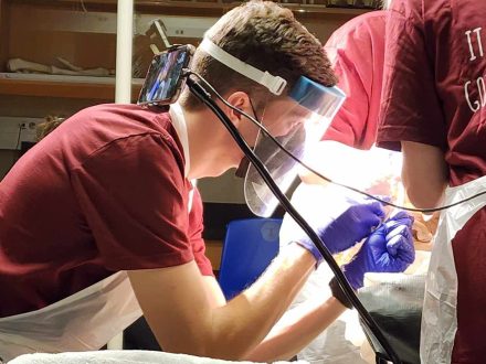 A student wearing a face shield and gloves performs a dissection in a lab setting, surrounded by surgical instruments laid out on a tray and assisted by others in matching maroon shirts.