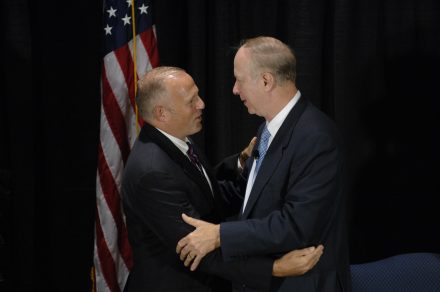 Two men on a stage shake hands and embrace warmly. They are wearing suits. An American flag is seen in the background.