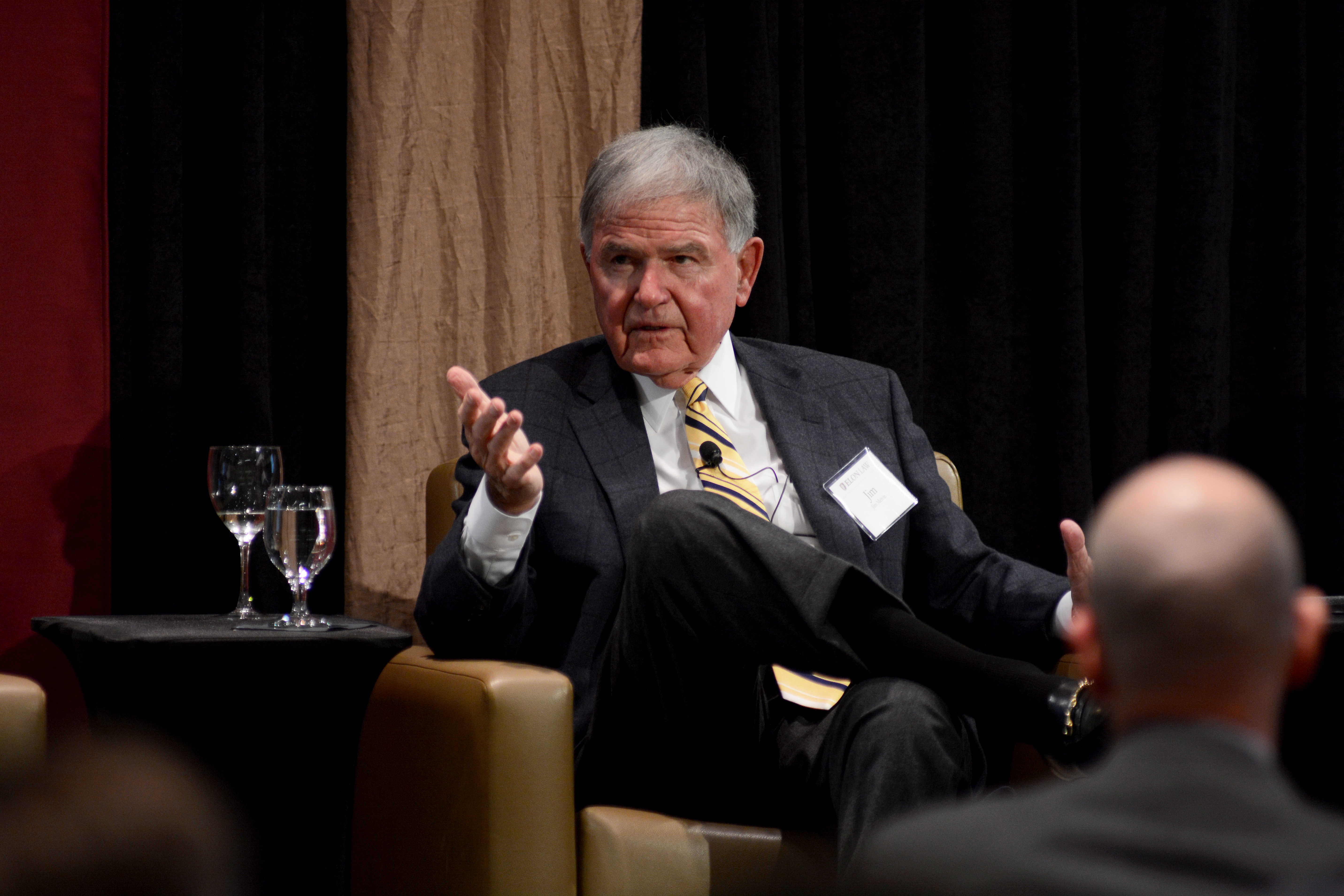 Jim Melvin in a suit, seated on a stage. He is gesturing while he speaks to an audience.