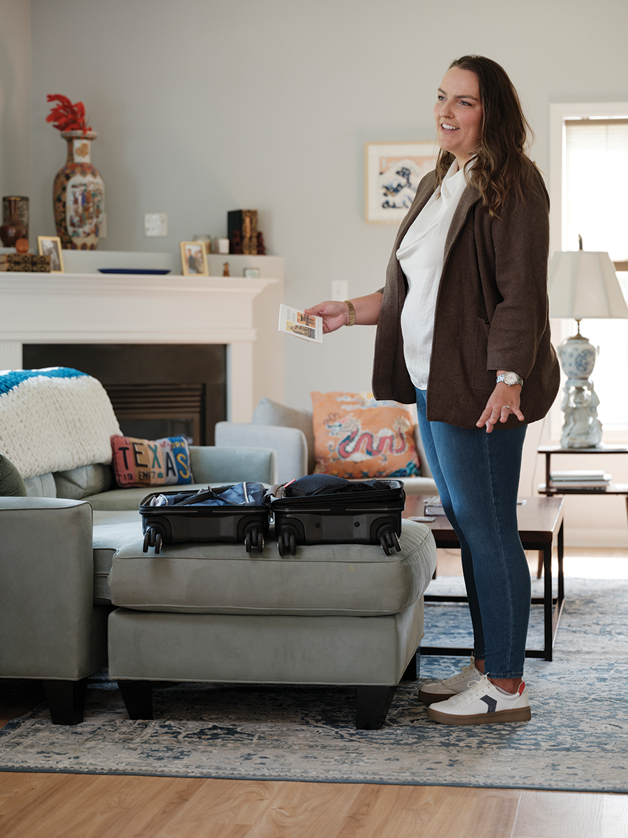 A woman stands next to a sofa and looks at a postcard. An open suitcase is on the sofa.