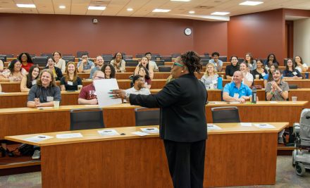 A professor standing in front of a crowded classroom. Students are seen laughing and smiling