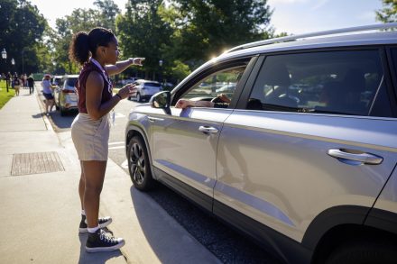 A young woman directs a driver in a silver SUV, pointing ahead while standing on a sunlit street lined with cars and trees.