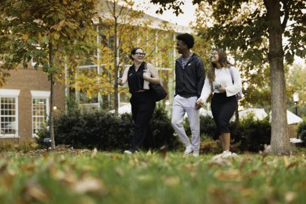 Three students walk together on a leafy campus path in autumn, smiling and talking with fall colors and brick buildings in the background.