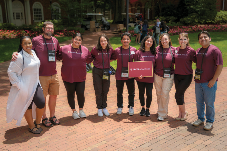 A group of Elon University employees pose together with their arms around each other on a brick walkway. They wear T-shirts that read "Elon Academy" and one woman holds an Elon Academy sign.