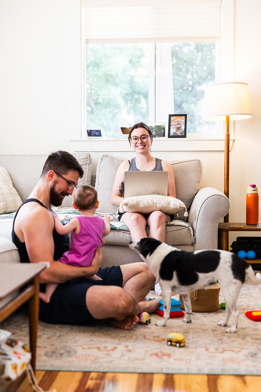 A man sits on the floor of a living room smiling at the baby he is holding in his lap. A dog stands beside him. A woman sits on a sofa nearby and looks up from the laptop in her lap, smiling.