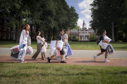 Students and families carry bags and bedding across campus during college move-in day, with a red-brick building in the background.