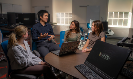 A professor and three students sit around a laptop and engage in discussion.