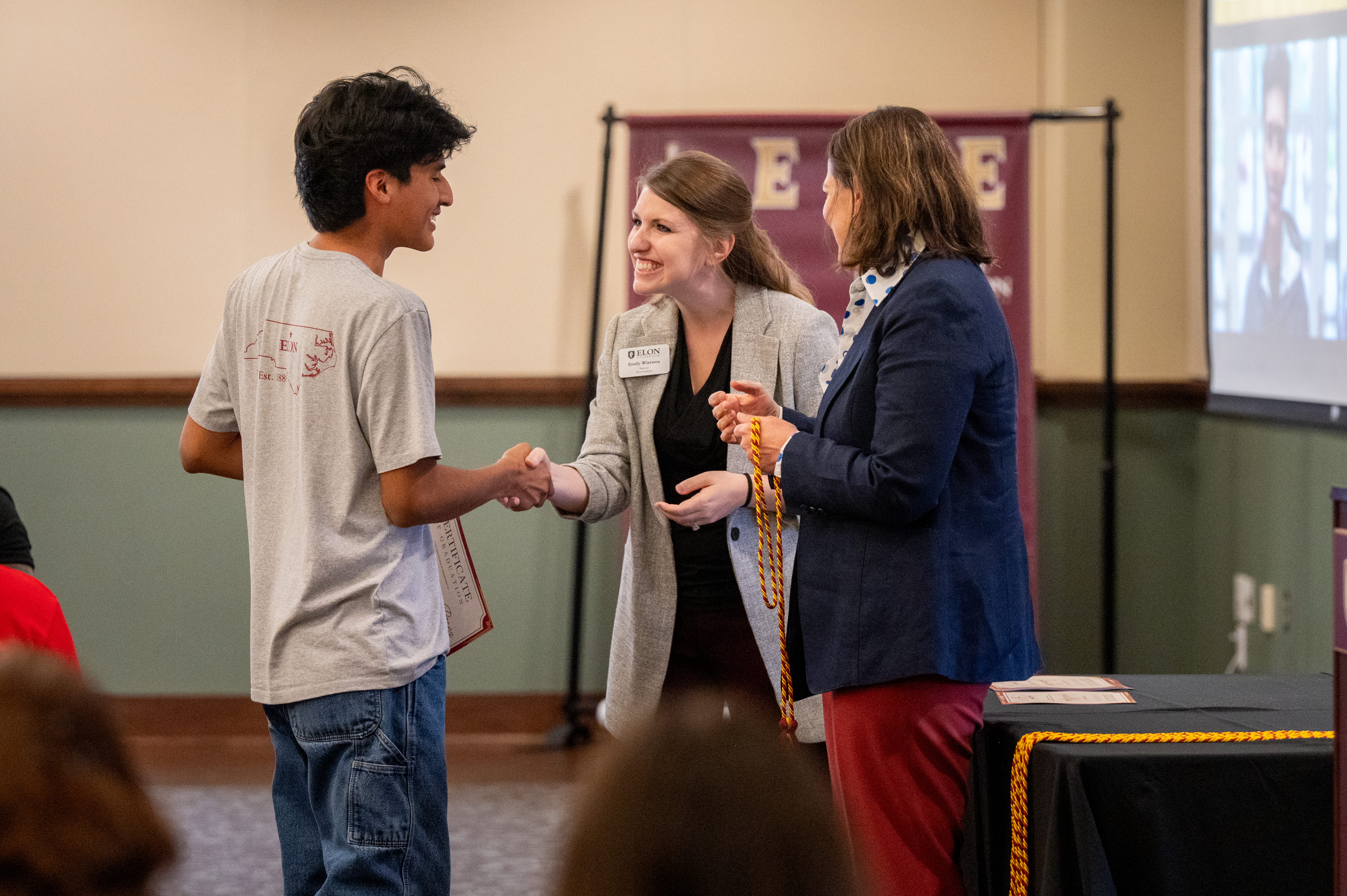 A student shakes hands with an Elon staff member while receiving a certificate, as President Connie Ledoux Book holds honor cords nearby.