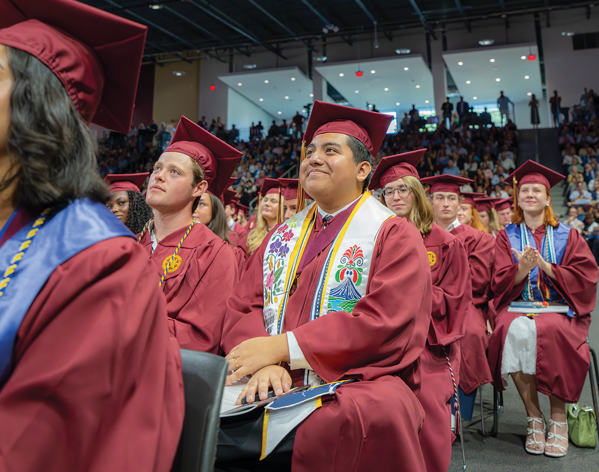 A male college graduate wearing a maroon cap and gown sits with a crowd of other graduates during a commencement ceremony and smiles.
