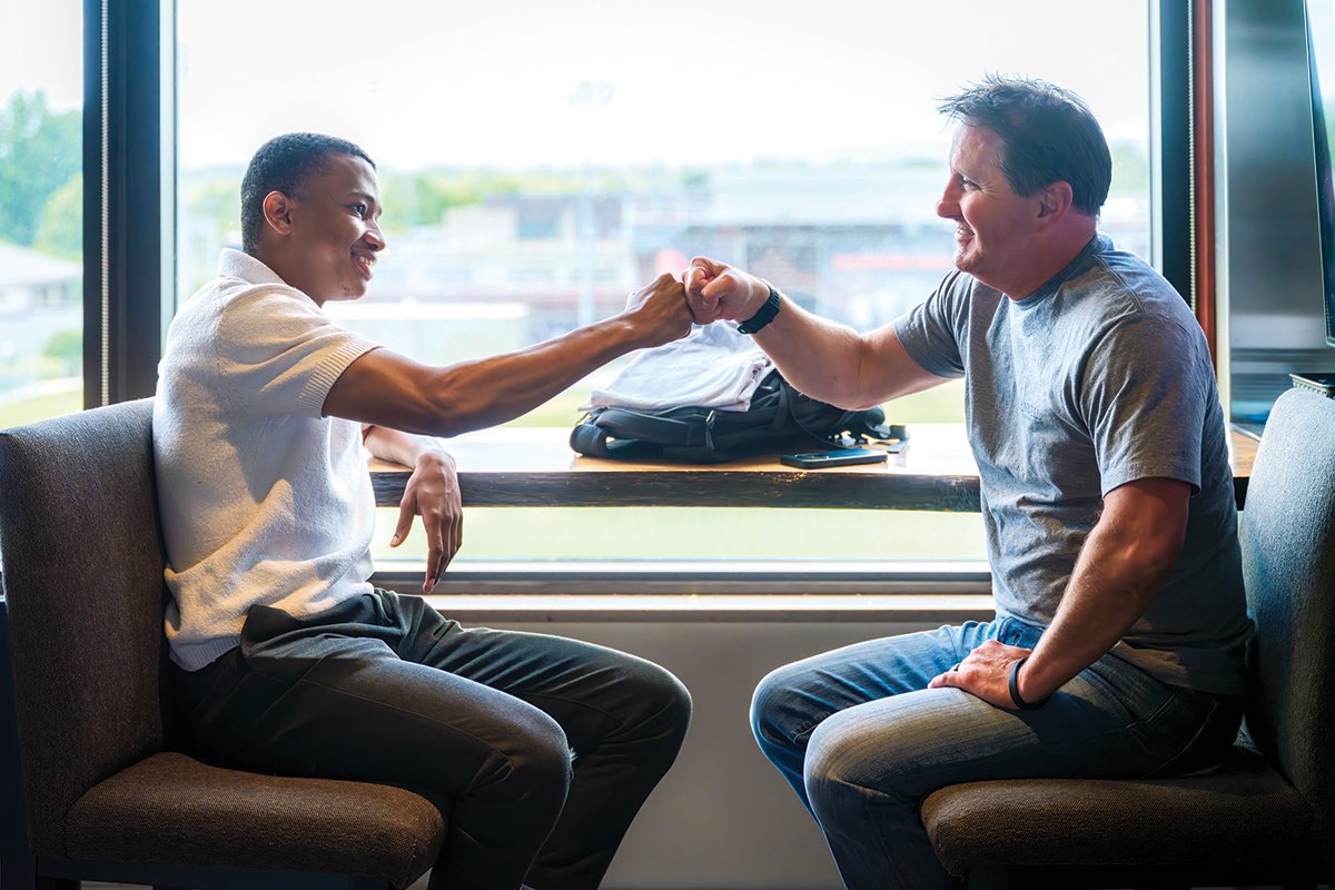Two men sit across from each other beside a table and give each other a fist bump.