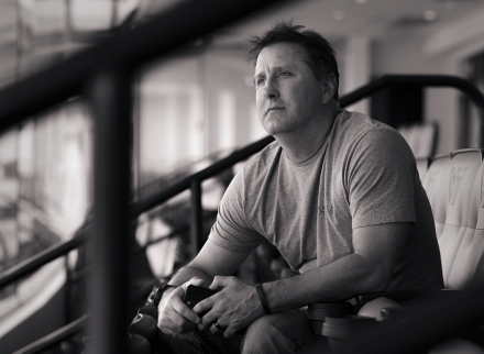 A black and white photo of a man sitting in a leather stadium seat, looking out across the distance to his right