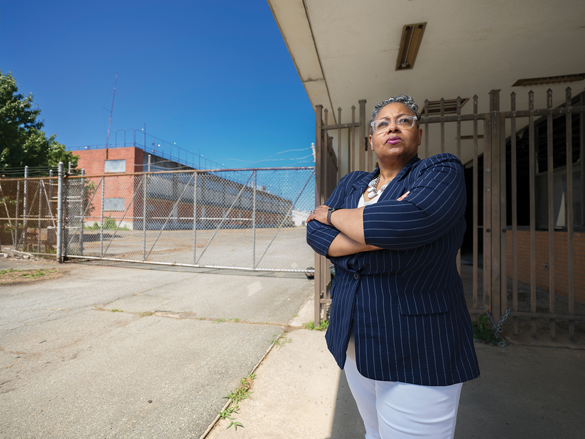 A woman stands purposefully with her head held high and her arms crossed over her chest outside of a run-down old building with a chain link fence in front of it.