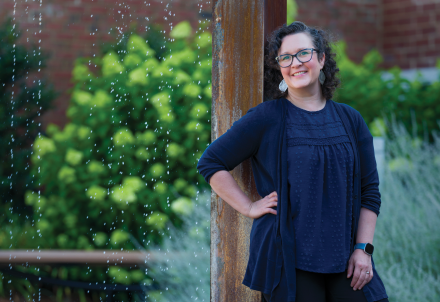 A woman poses leaned against a pillar with her hand on her hip as water from a fountain cascades down behind her.