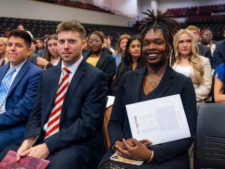 Several rows of students in a crowded auditorium. The front row is shown smiling at the camera.