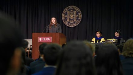A woman at a podium on a stage. Administrators in academic regalia are seated on the stage nearby.