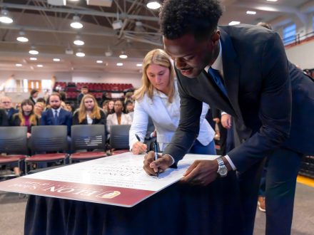 A man in a suit signs his name on a maroon and white poster. A woman is shown behind him signing another part of the poster.