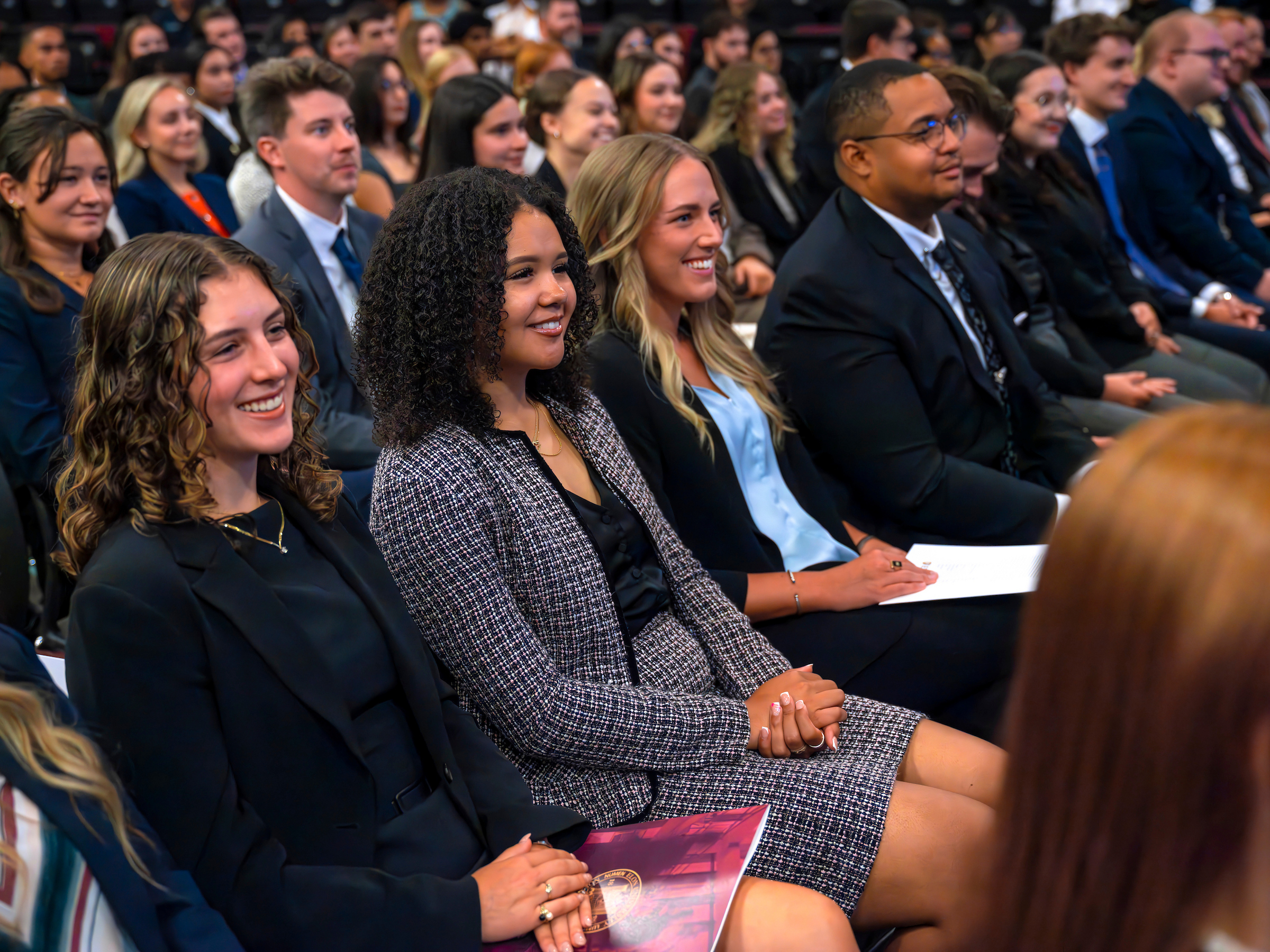 A large group of professionally dressed law students seated in an auditorium. They are smiling and laughing at a speaker's remarks.