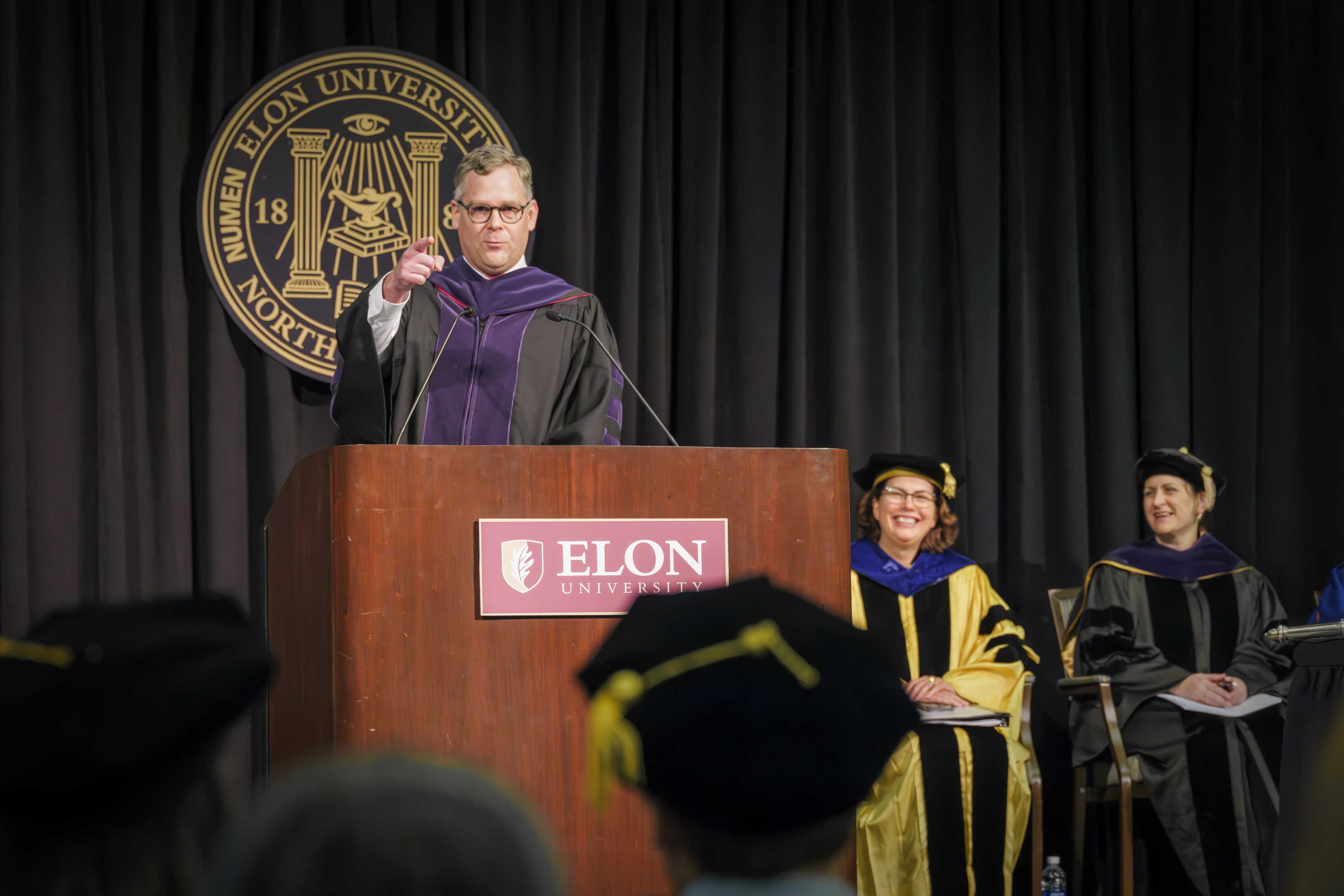 A man speaks at an Elon University podium, gesturing toward the audience. People in academic regalia are seated on the stage to the right.