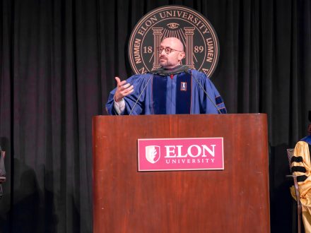 A man at a podium in academic regalia gestures while speaking. He is on a stge with Elon University's seal behind him.