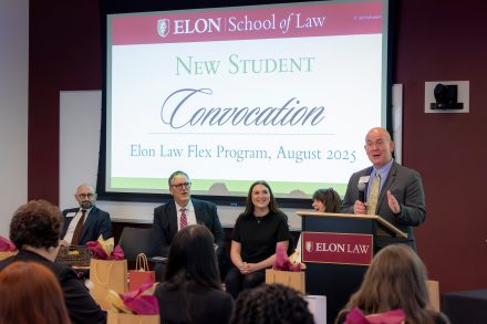 A man at a podium with an Elon Law plaquard and a screen that says New Student Convocation, Elon Law Flex Program, August 2025 projected behind him. Several are seated to his left, smiling and laughing.