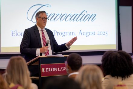Frank Whitney in a suit and tie, speaking into a microphone behind a podium with an "Elon Law" plaquard on it. The word "Convocation" is projected on screen behind him in decorative script.